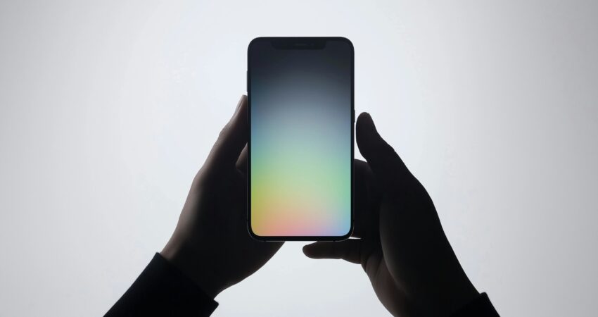 The image depicts a pair of hands holding a smartphone against a plain, light background. The phone's screen displays a gradient of soft colors, transitioning from blue at the top to pink at the bottom, creating a calming visual effect. The hands are silhouetted, emphasizing the phone as the central subject. The simplicity of the composition draws attention to the sleek design of the device, highlighting its modern and minimalist aesthetic. The interaction between the hands and the phone suggests a focus on technology and connectivity. The overall tone of the image is serene and futuristic, with the gradient colors adding a touch of elegance.