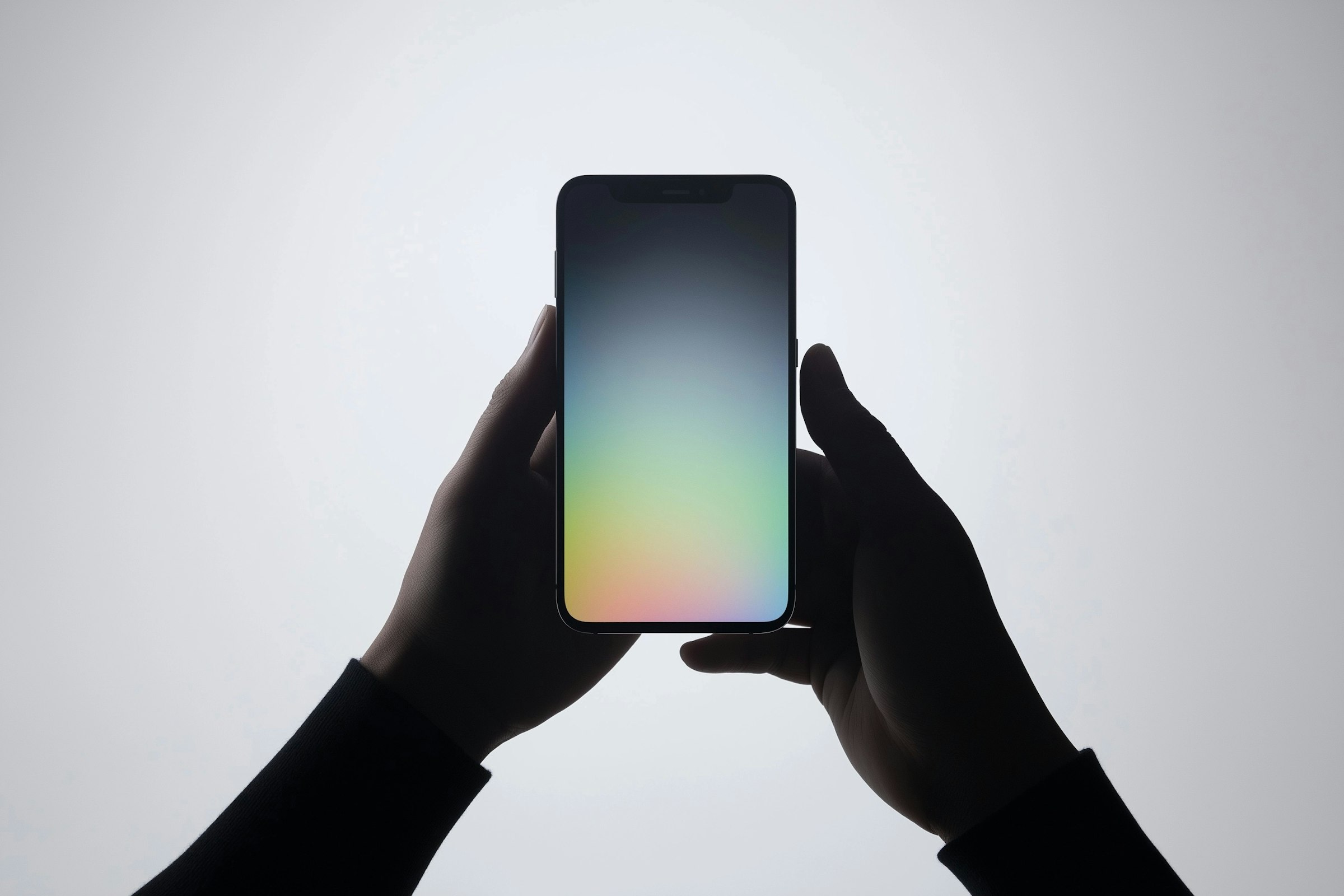 The image depicts a pair of hands holding a smartphone against a plain, light background. The phone's screen displays a gradient of soft colors, transitioning from blue at the top to pink at the bottom, creating a calming visual effect. The hands are silhouetted, emphasizing the phone as the central subject. The simplicity of the composition draws attention to the sleek design of the device, highlighting its modern and minimalist aesthetic. The interaction between the hands and the phone suggests a focus on technology and connectivity. The overall tone of the image is serene and futuristic, with the gradient colors adding a touch of elegance.